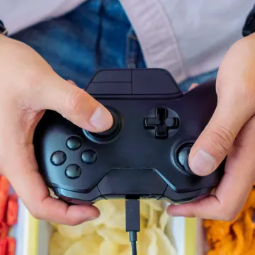 © Paolaulloa0022 / AdobeStock Person hält einen Gamecontroller über Schalen mit Snacks